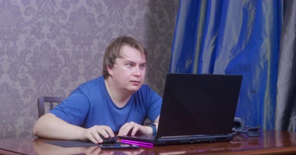 Młody człowiek w niebieskiej koszulce z głupim spojrzeniem siedzi przy stole w biurze, używa laptopa i myszki komputerowej, dłubie w nosie, a potem je gluty. Obrzydliwe nawyki. — Wideo stockowe