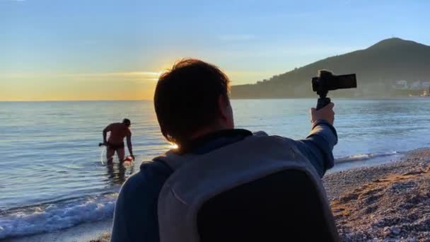 Чоловік сидить на пляжі і знімається на камеру, розповідаючи щось під час заходу сонця. Інший хлопець в цей час виходить з води, несучи дивний дизайн водолазної трубки, шланга і пластикової пляшки . — стокове відео