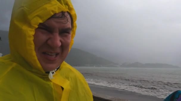 Junger Mann in gelbem Regenmantel verlässt bei schlechtem Wetter das Haus: Es regnet, starker Wind bläst ihm ins Gesicht, und im Hintergrund ist die See stürmisch. Guy versucht etwas zu erzählen und gestikuliert emotional. — Stockvideo