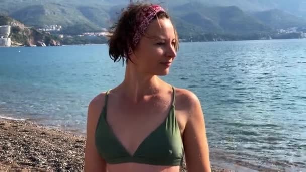 Mujer atractiva joven con una banda de tela en la cabeza y en traje de baño de dos piezas posa en la playa, el mar y el paisaje de montaña en el fondo. — Vídeos de Stock