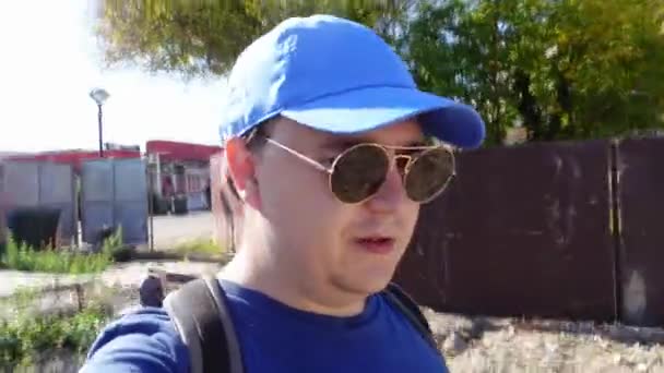 Mężczyzna w czapce baseballowej i okularach przeciera się przez nowy obszar w trakcie budowy i z entuzjazmem o czymś mówi. Travel blogger sprawia, że raport o życiu innego odwiedzanego miejsca. — Wideo stockowe