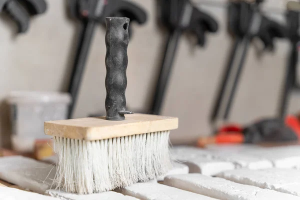 Großer Pinsel, mit dem Handwerker früher die Oberfläche von dekorativen Fliesen vom Staub reinigten, steht auf dem Schreibtisch im Vordergrund, Nahaufnahme, Seitenansicht. Andere Arbeitsgeräte und Handschuhe im unscharfen Hintergrund. Stockbild