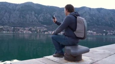 Turist sırt çantalı genç adam, dağların eteklerindeki sahil ve sahil şeridinde çekim yapıyor, aksiyon kamerası kullanıyor. Seyahat blogcusu vlog 'u için içerik oluşturdu