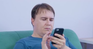 Kısa sarı saçlı, olgun, tombul bir adam internette haber okuyor. Akıllı telefonuyla. Oturma odasındaki yumuşak koltukta oturuyor.
