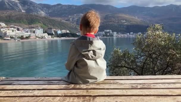 身穿外套的男孩悲伤地坐在长椅上 欣赏美丽的海景 沐浴在阳光下 他被父母冒犯了 所以当有人喊他时 他转过身去 立刻转身离去 — 图库视频影像
