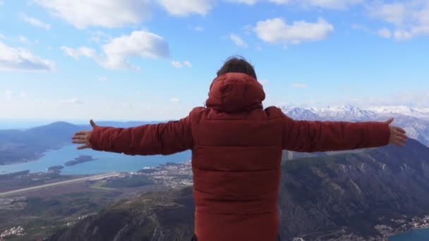 Persona con chaqueta hinchable se encuentra en la cima de la montaña y admira el hermoso paisaje con cordillera y bahía, extendiendo los brazos a los lados, sintiendo libertad, vista desde la parte posterior. — Vídeo de stock