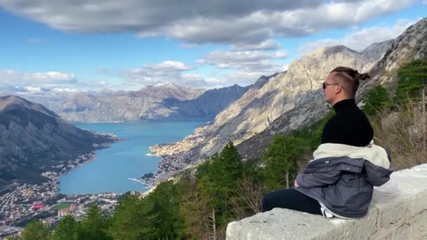 Jongeman met bril en stijlvolle kapsel zit op bakstenen borstwering en bewondert prachtige baai, uitzicht vanaf de hoogte van de bergen. Sfeer van vrede en rust. — Stockvideo