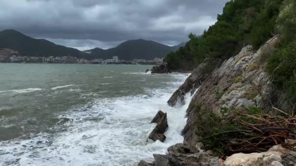 Hullámok csapódnak egy növényzettel borított sziklába, a magasból nézve. Durva szürke tengerpart viharos tengerrel és üdülővárossal a hegyek lábánál a felhős égbolttal szemben. — Stock videók