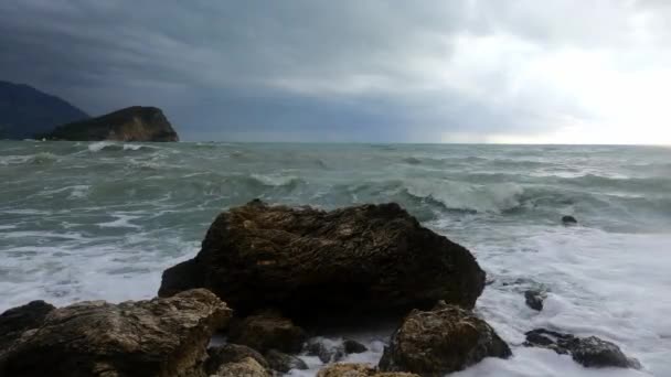 Onde scatenate rotolano su grandi rocce sulla costa, tagliando l'acqua come frangiflutti. Tempo nuvoloso e mare inquieto dopo o prima di una tempesta, vista dalla riva. — Video Stock