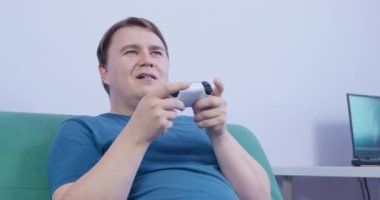 Dişlerinde diş teli olan ve tatmin olmuş bir şekilde oyun konsolunun kontrolünü elinde tutan genç bir adam, evinde kanepede, ön tarafta ilginç bir video oyunu oynuyor..