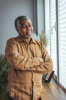 Mutlu bir Afrikalı emeklinin yan görüntüsü. Pencere kenarında oturan yaşlı bir adam. Tatmin olmuş yaşlı bir adam pencereden dışarı bakar ve pencerenin yanında dururken gülümser. Olumlu ve kendinden emin yaşlılar emekliliğin tadını çıkarıyor.