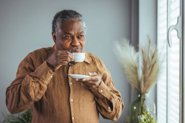 Yaşlı Afrikalı bir adam sabahları pencerenin önünde durmuş kahve içerken mutlu, yaşlı bir Afrikalı adam elinde bir fincan kahveyle evde, emekli, yaşlı ve sağlıklı bir hayat sürüyor. Emeklilik yaşam tarzı, evde zaman geçirme.