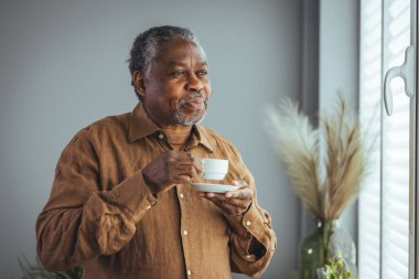 Yaşlı Afrikalı bir adam sabahları pencerenin önünde durmuş kahve içerken mutlu, yaşlı bir Afrikalı adam elinde bir fincan kahveyle evde, emekli, yaşlı ve sağlıklı bir hayat sürüyor. Emeklilik yaşam tarzı, evde zaman geçirme.