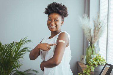 Aşılanmış Afrikalı Amerikalı genç bir kadın kovid 19 aşısından sonra kollarını gösteriyor, gri arka planda duruyor, içi boş. Corona virüsü kavramını durdur