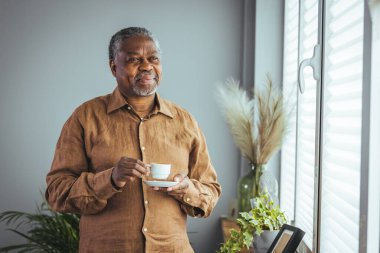 Yaşlı Afrikalı bir adam sabahları pencerenin önünde durmuş kahve içerken mutlu, yaşlı bir Afrikalı adam elinde bir fincan kahveyle evde, emekli, yaşlı ve sağlıklı bir hayat sürüyor. Emeklilik yaşam tarzı, evde zaman geçirme.