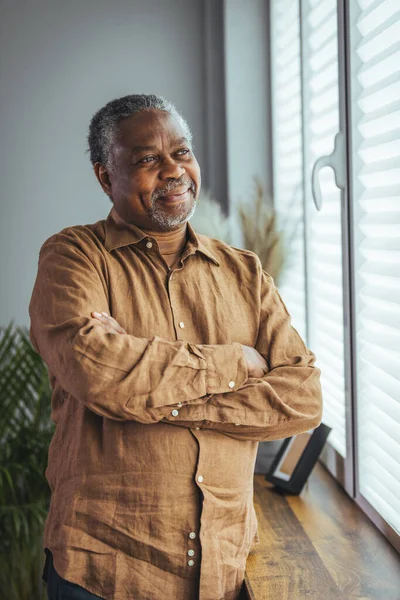 Side view of a happy African retired elderly man standing at home by the window. A satisfied old man looks out the window and smiles as he stands by the window. Positive and self-confident seniors enjoy retirement.