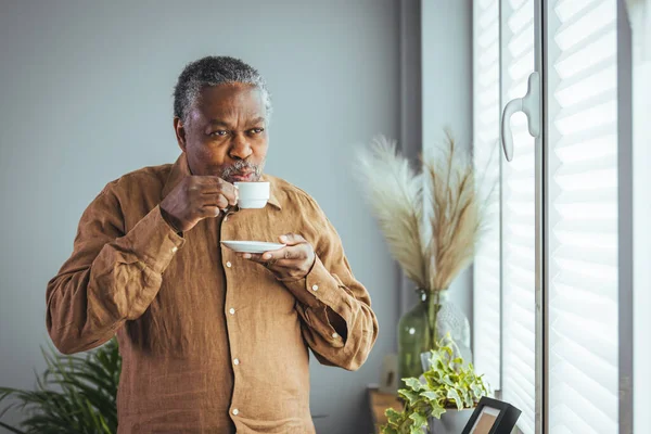 Older African man drinking coffee while standing by the window in the morning, happy older African man holding a cup of coffee at home, retired old people healthy lifestyle. Retirement lifestyle, spending time at home