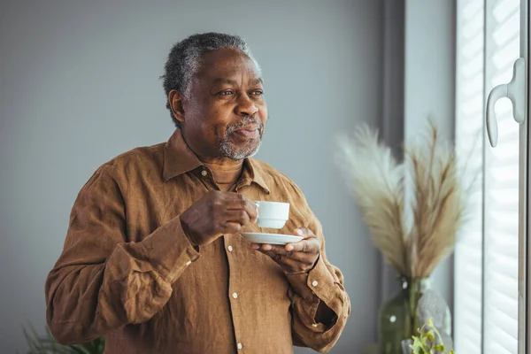 Older African man drinking coffee while standing by the window in the morning, happy older African man holding a cup of coffee at home, retired old people healthy lifestyle. Retirement lifestyle, spending time at home