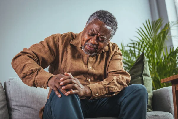 Photo of mature, elderly man sitting on a sofa in the living room at home and touching his knee by the pain during the day. Mature man massaging his painful knee.