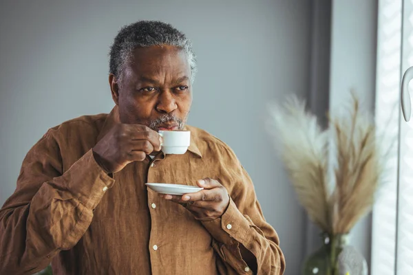 彼の手にコーヒーの白いカップを持つ孤独な引退したアフリカの老人は窓の外を見て 隔離中にガラスの後ろに立っています 概念コロナウイルスパンデミック — ストック写真