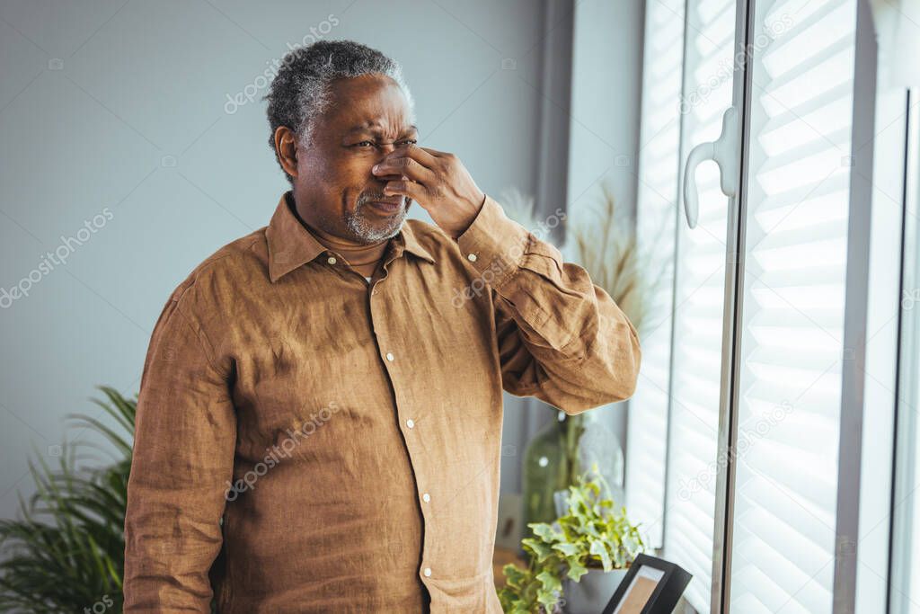 A tired, depressed older African man is sitting on the couch in the living room, feeling hurt and lonely. An older, gray-haired man touches his head, suffers from a severe headache or remembers bad memories