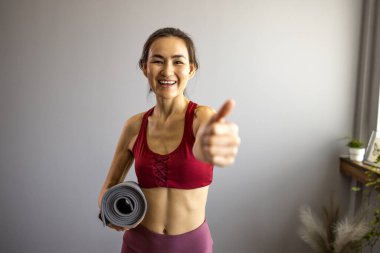 Yoga minderi tutan mutlu Asyalı kız ve kamera u dnevnoj sobi izliyor. Gülümseyen kadın spor yapmak için sabırsızlanıyor ve yoğun bir iş gününden sonra spor salonuna gitmeye hazırlanıyor. Parmağını kaldırdı.