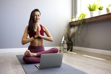 Asyalı genç bir spor kadını oturma odasında yerde oturmuş online bilgisayar dersinde yoga yapıyor. Sakin bir kadın dizüstü bilgisayarında web kamerasıyla dersleri izlerken evde meditasyon yapıyor. Rahatlama ve yoga kavramı.