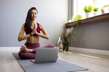 Asyalı genç bir spor kadını oturma odasında yerde oturmuş online bilgisayar dersinde yoga yapıyor. Sakin bir kadın dizüstü bilgisayarında web kamerasıyla dersleri izlerken evde meditasyon yapıyor. Rahatlama ve yoga kavramı.