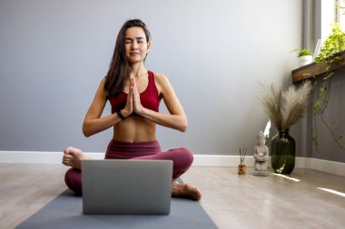 Evde dizüstü bilgisayar başında yoga egzersizi yapan zinde bir kadın. Çekici Çinli kadın paspasın üzerinde egzersiz yapıyor ve dizüstü bilgisayarda eğitici videolar izliyor..