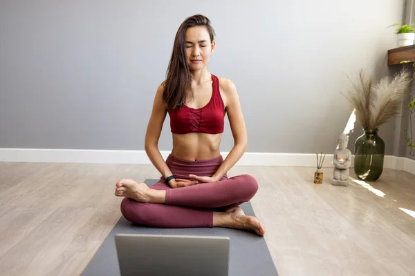 An Asian young sports woman is sitting on the floor in the living room, practicing yoga in an online computer class. A calm woman meditates at home watching classes on a webcam on a laptop. The concept of relaxation and yoga