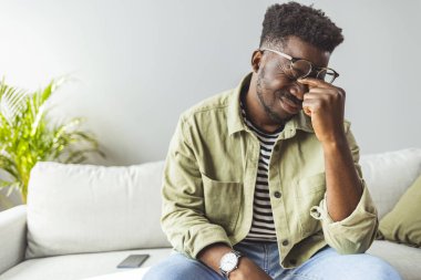 Genç Afrikalı adam elleriyle kafasına dokunuyor ve evdeki kanepede otururken gözlerini kapalı tutuyor. Zenci adam stresli ve baş ağrısı çekiyor. Hasta ve yorgun hissediyorum.. 