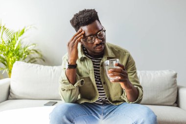 Evde stresli görünen bir gencin fotoğrafı. Hüsrana uğramış yakışıklı genç adam başını okşuyor ve evdeki kanepede otururken gözlerini kapalı tutuyor.