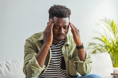 Evdeki kanepede otururken stresli görünen bir adamın fotoğrafı. Zor bir günün ardından baş ağrısı çeken genç Afro-Amerikalı iş adamı, evdeki kanepede oturup, uzayı kopyalıyor.