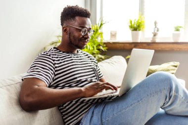 Kanepede dizüstü bilgisayarlı Afro-Amerikan bir adam oturuyor.  