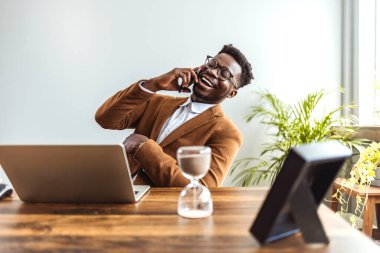 Afro-Amerikan iş adamı laptopuyla ve kum saatiyle masada oturuyor ve akıllı telefondan konuşuyor.