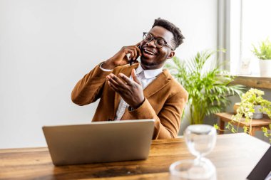 Afro-Amerikan iş adamı laptopuyla ve kum saatiyle masada oturuyor ve akıllı telefondan konuşuyor.