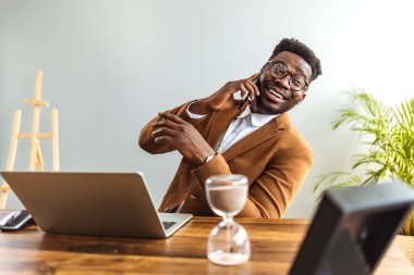 african american businessman sitting at table with laptop and hourglass and talking on smartphone