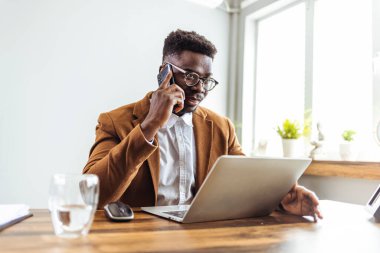 Kendine güvenen Afrikalı Amerikalı iş adamı dizüstü bilgisayarla oturmuş telefonla konuşuyor. 