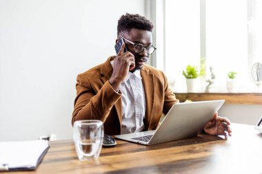 Kendine güvenen Afrikalı Amerikalı iş adamı dizüstü bilgisayarla oturmuş telefonla konuşuyor. 