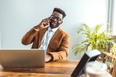 Kendine güvenen Afrikalı Amerikalı iş adamı dizüstü bilgisayarla oturmuş telefonla konuşuyor. 