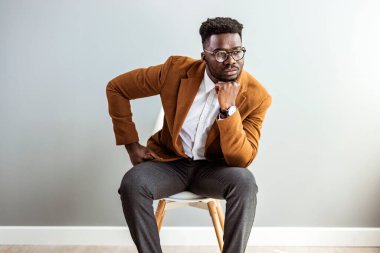Genç Afro-Amerikalı adam, elleri çenesinde gri duvar arkasında, sandalyede oturuyor.