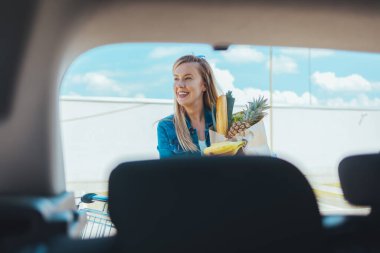 Sokakta alışveriş torbası taşıyan genç bir kadın. Arabanın ön camından görüntüle
