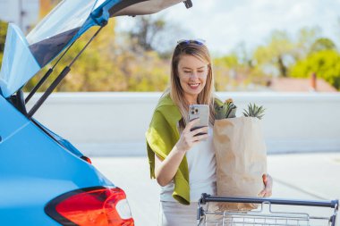 Alışveriş çantalı genç bir kadın arabanın yanında akıllı telefon kullanıyor.
