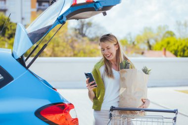 Alışveriş çantalı genç bir kadın arabanın yanında akıllı telefon kullanıyor.