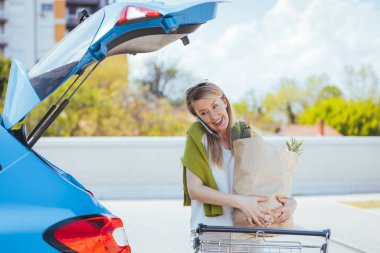 Sokakta alışveriş torbası taşıyan genç bir kadın cep telefonuyla konuşuyor.