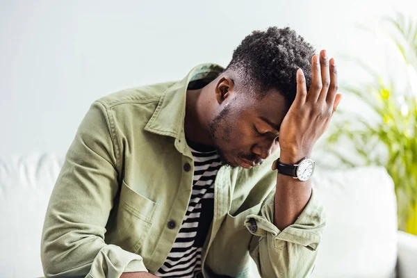 Evde stresli görünen bir gencin fotoğrafı. Hüsrana uğramış yakışıklı genç adam başını okşuyor ve evdeki kanepede otururken gözlerini kapalı tutuyor.