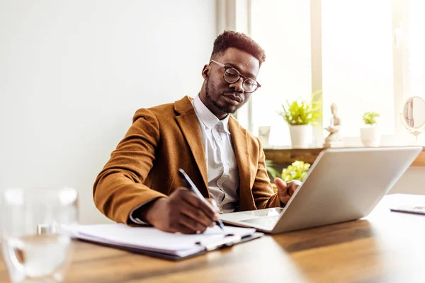 自宅で働いている若いエレガントな男 アフリカの深刻なビジネスマンノートを書き ラップトップを使用しています 仕事中にデスクに座っている黒人起業家に焦点を当てた — ストック写真