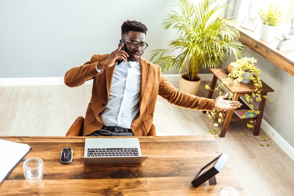 Kendine güvenen Afrikalı Amerikalı iş adamı dizüstü bilgisayarla oturmuş telefonla konuşuyor. 