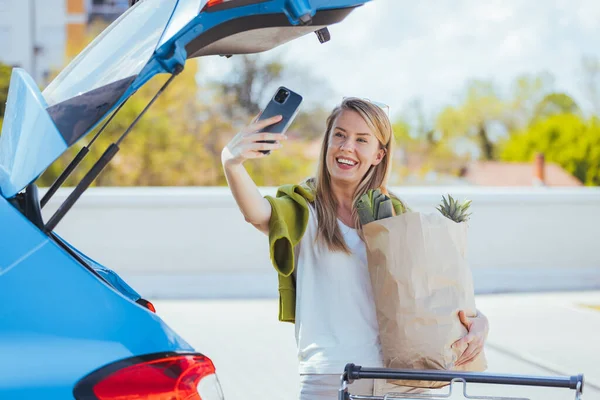 買い物袋を持った若い女性が車の近くのスマートフォンで自撮り — ストック写真
