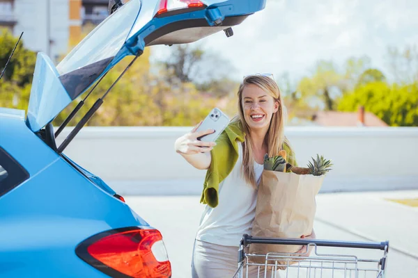 買い物袋を持った若い女性が車の近くのスマートフォンで自撮り — ストック写真